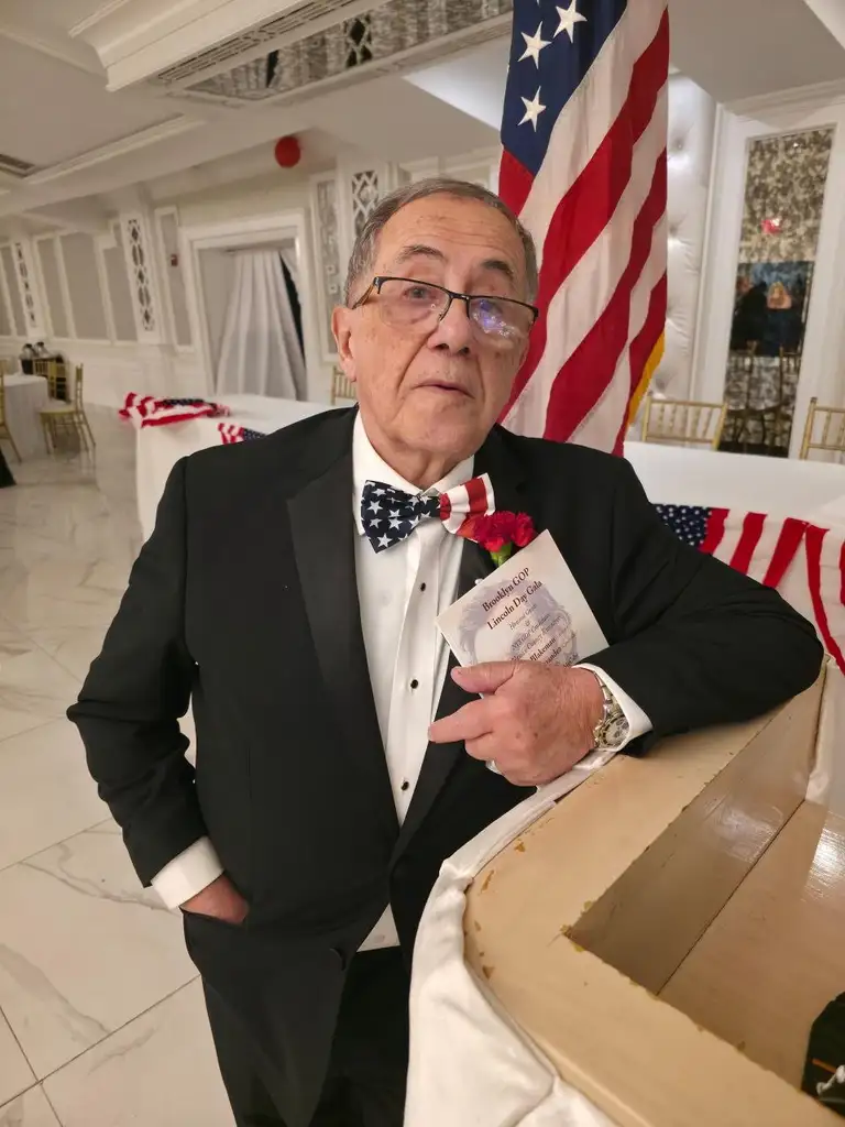 A guest in a stars-and-stripes bow tie holds a Brooklyn GOP Lincoln Day Gala program beside an American flag